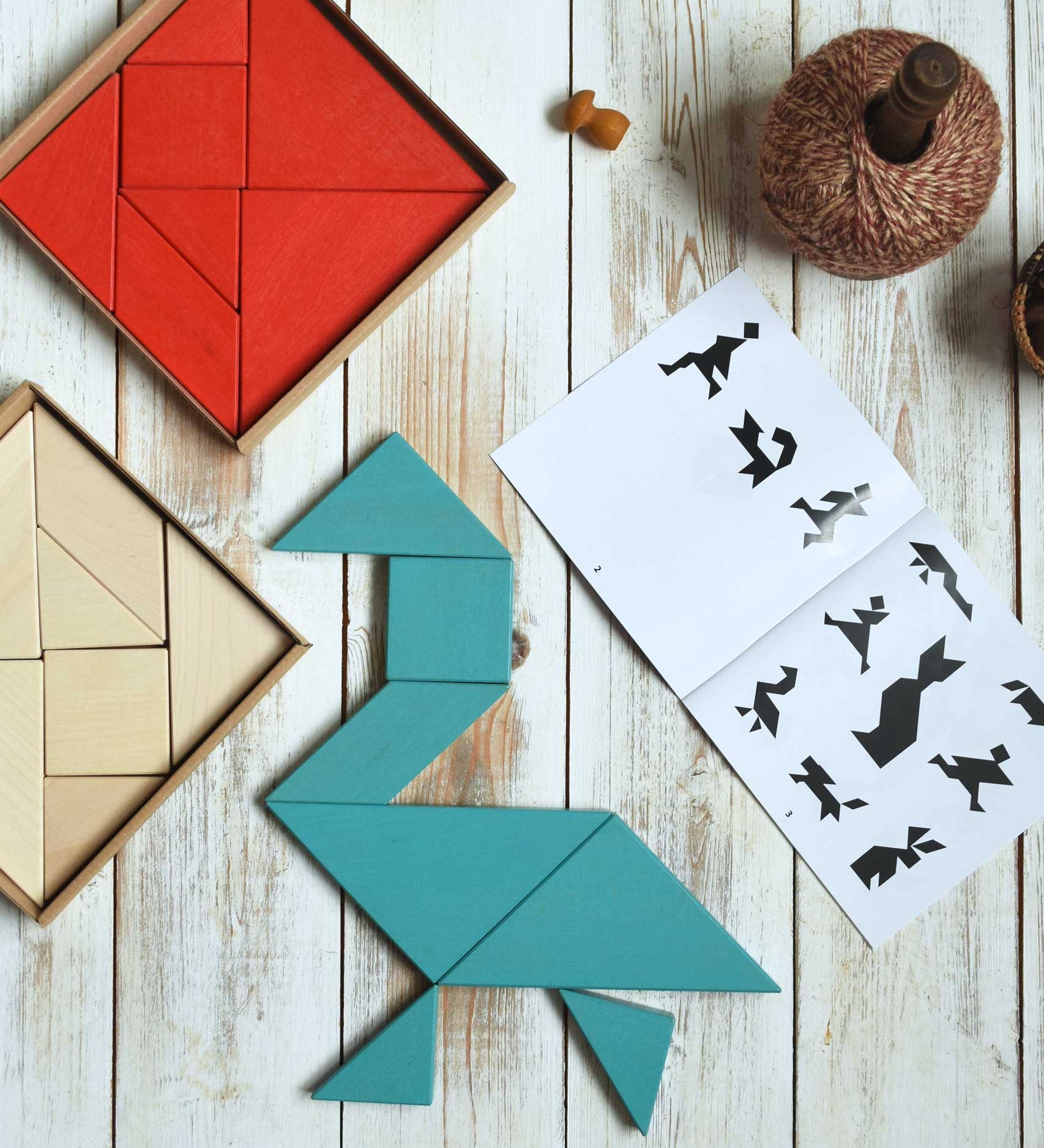 A blue Bajo Tangram puzzle placed on a white wooden surface. The puzzle has been placed in the shape of a bird. The accompanying ideas booklet can be seen open besides it with the red and natural tangram puzzle placed the other side. These heirloom quality toys by Bajo are part of a wide range of wooden toys available here at Babipur. 