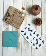 The blue Bajo Tangram wooden toy puzzle placed on a white wooden surface. The pieces of the puzzle have been placed in a square shape next to the packaging and accompanying ideas booklet. These heirloom quality toys by Bajo are part of a wide range of wooden toys available here at Babipur. 