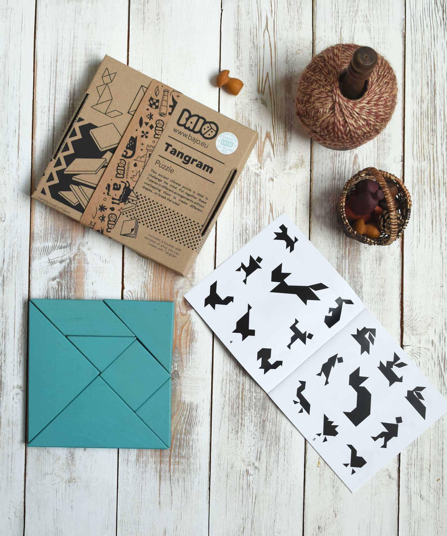 The blue Bajo Tangram wooden toy puzzle placed on a white wooden surface. The pieces of the puzzle have been placed in a square shape next to the packaging and accompanying ideas booklet. These heirloom quality toys by Bajo are part of a wide range of wooden toys available here at Babipur. 