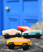 Bajo yellow ToBe Poly car wooden toy on a stone wall with other colours in the ebackground.