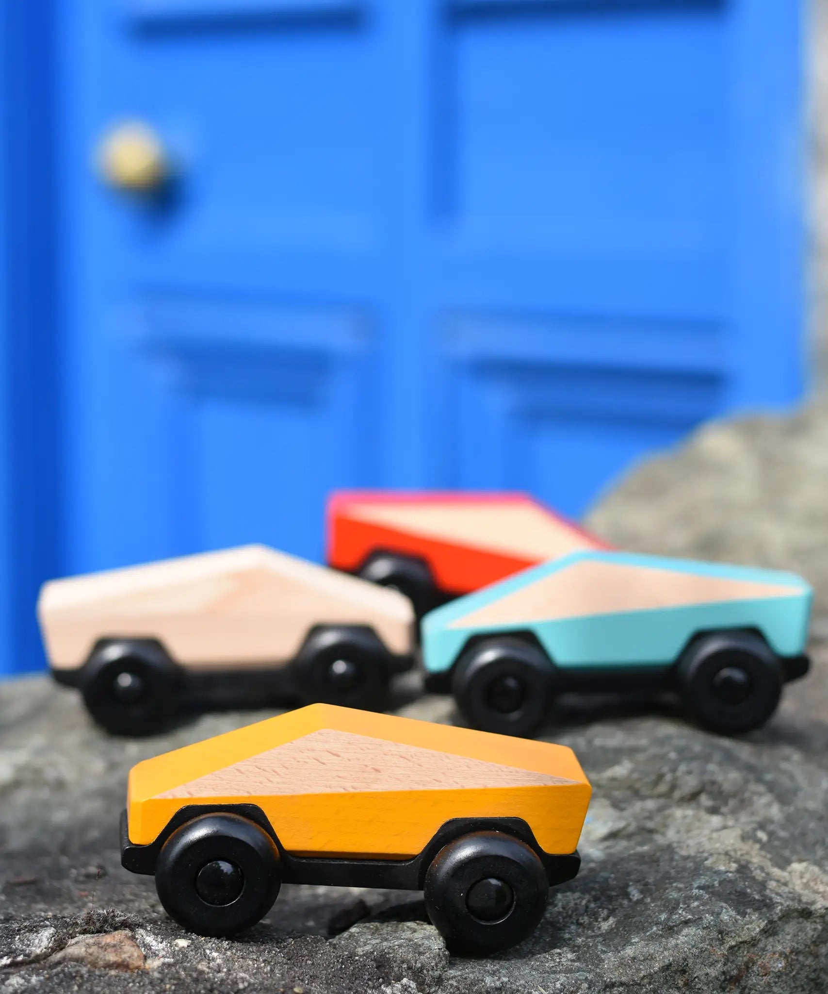 Bajo yellow ToBe Poly car wooden toy on a stone wall with other colours in the ebackground.