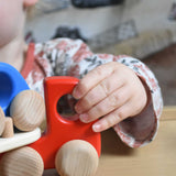 A close up of a child's hand holding on the front of the red coloured Bajo Auto Transporter. The wooden toy car transporter comes with two cars, one green and one blue. These heirloom quality toys by Bajo are part of a wide range of wooden toys available here at Babipur. 