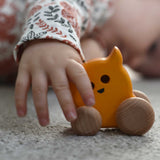 A child's hand holding onto a Bajo push along eye ghost wooden toy. The toy has a yellow painted body and natural wooden wheels. These heirloom quality toys by Bajo are part of a wide range of wooden toys available here at Babipur. 