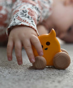 A child's hand holding onto a Bajo push along eye ghost wooden toy. The toy has a yellow painted body and natural wooden wheels. These heirloom quality toys by Bajo are part of a wide range of wooden toys available here at Babipur. 