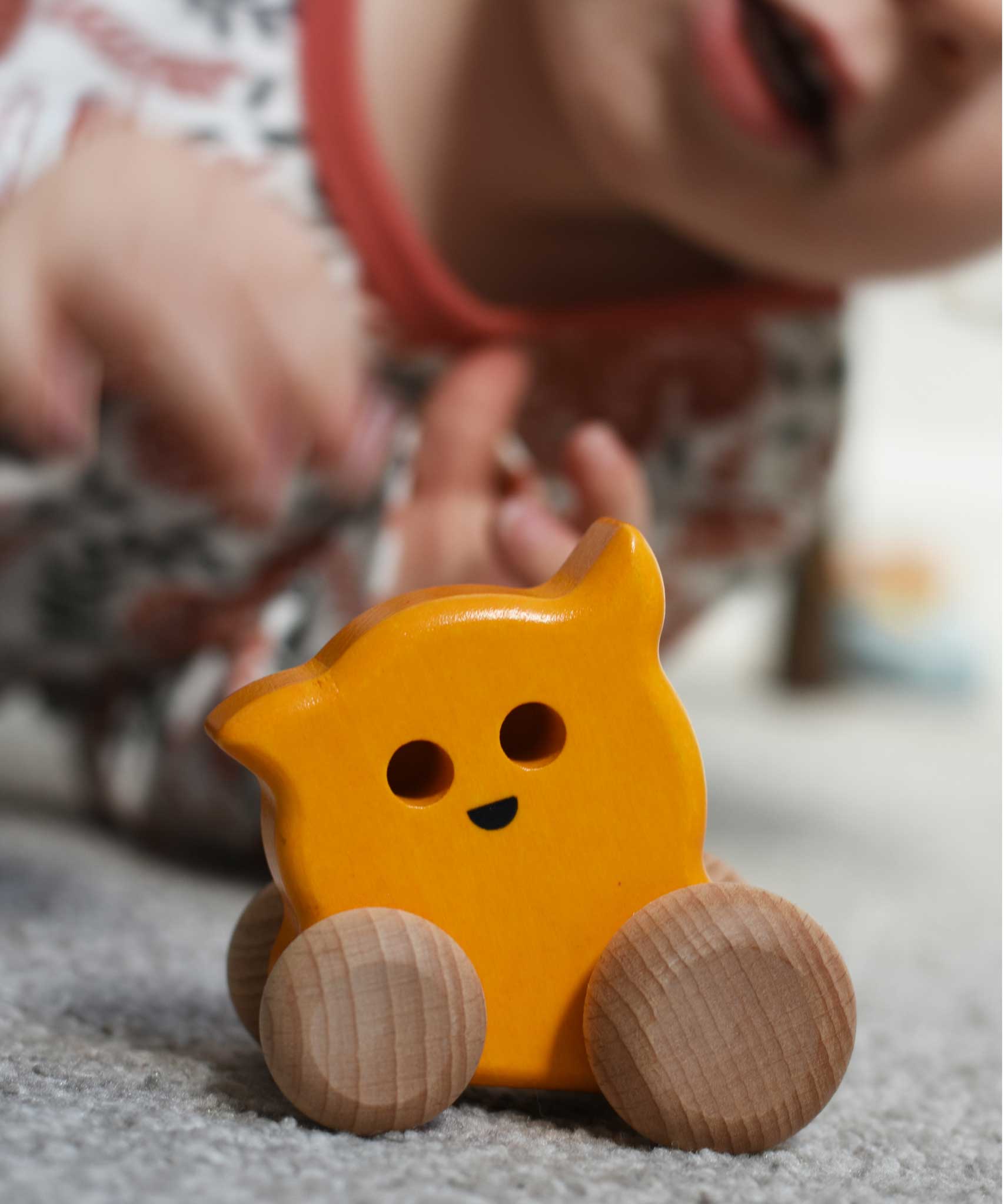 A Bajo push along eye ghost wooden toy on carpet, a child can be seen in the background. The toy has a yellow painted body and natural wooden wheels. These heirloom quality toys by Bajo are part of a wide range of wooden toys available here at Babipur. 