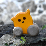 A Bajo push along eye ghost wooden toy placed on a stone wall outdoors. Toe toy has a yellow painted body and natural wooden wheels. These heirloom quality toys by Bajo are part of a wide range of wooden toys available here at Babipur. 