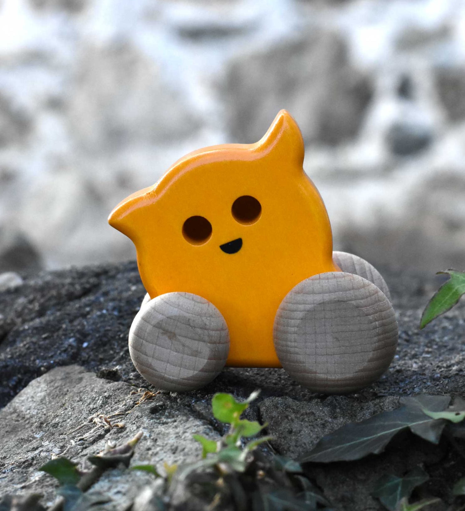 A Bajo push along eye ghost wooden toy placed on a stone wall outdoors. Toe toy has a yellow painted body and natural wooden wheels. These heirloom quality toys by Bajo are part of a wide range of wooden toys available here at Babipur. 