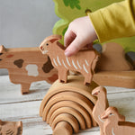 A child's hand holding the goat figure from the Bajo farm animals set above a mini Grimm's natural rainbow. Other animal figures can be seen in the background.
