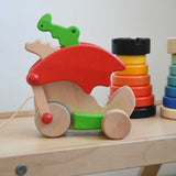 A Bajo green pull along dragon wooden toy placed on a Triclimb Miri slide. The dragon toy features two wheels at the front and back and red wing that move along with the front wheels. Other Bajo wooden toys can be seen in the background. These heirloom quality toys by Bajo are part of a wide range of wooden toys available here at Babipur.