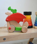 A Bajo green pull along dragon wooden toy placed on a Triclimb Miri slide. The dragon toy features two wheels at the front and back and red wing that move along with the front wheels. Other Bajo wooden toys can be seen in the background. These heirloom quality toys by Bajo are part of a wide range of wooden toys available here at Babipur.
