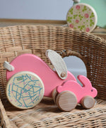 A small pink coloured Bajo rabbit placed on a wicker chair with a big pink rabbit behind it.  These heirloom quality toys by Bajo are part of a wide range of wooden toys available here at Babipur.