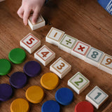 An overhead view of a child plaing with the Bajo 40 Numbers & Symbols Blocks. They have number blocks with Grapat wooden coins lined up besides them. These heirloom quality toys by Bajo are part of a wide range of wooden toys available here at Babipur. 