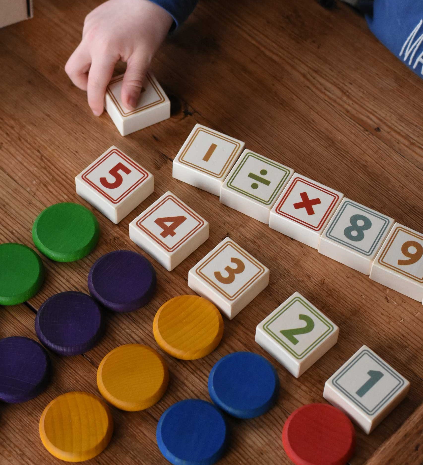 An overhead view of a child playing with the Bajo 40 Numbers & Symbols Blocks. They have number blocks with Grapat wooden coins lined up besides them. These heirloom quality toys by Bajo are part of a wide range of wooden toys available here at Babipur. 