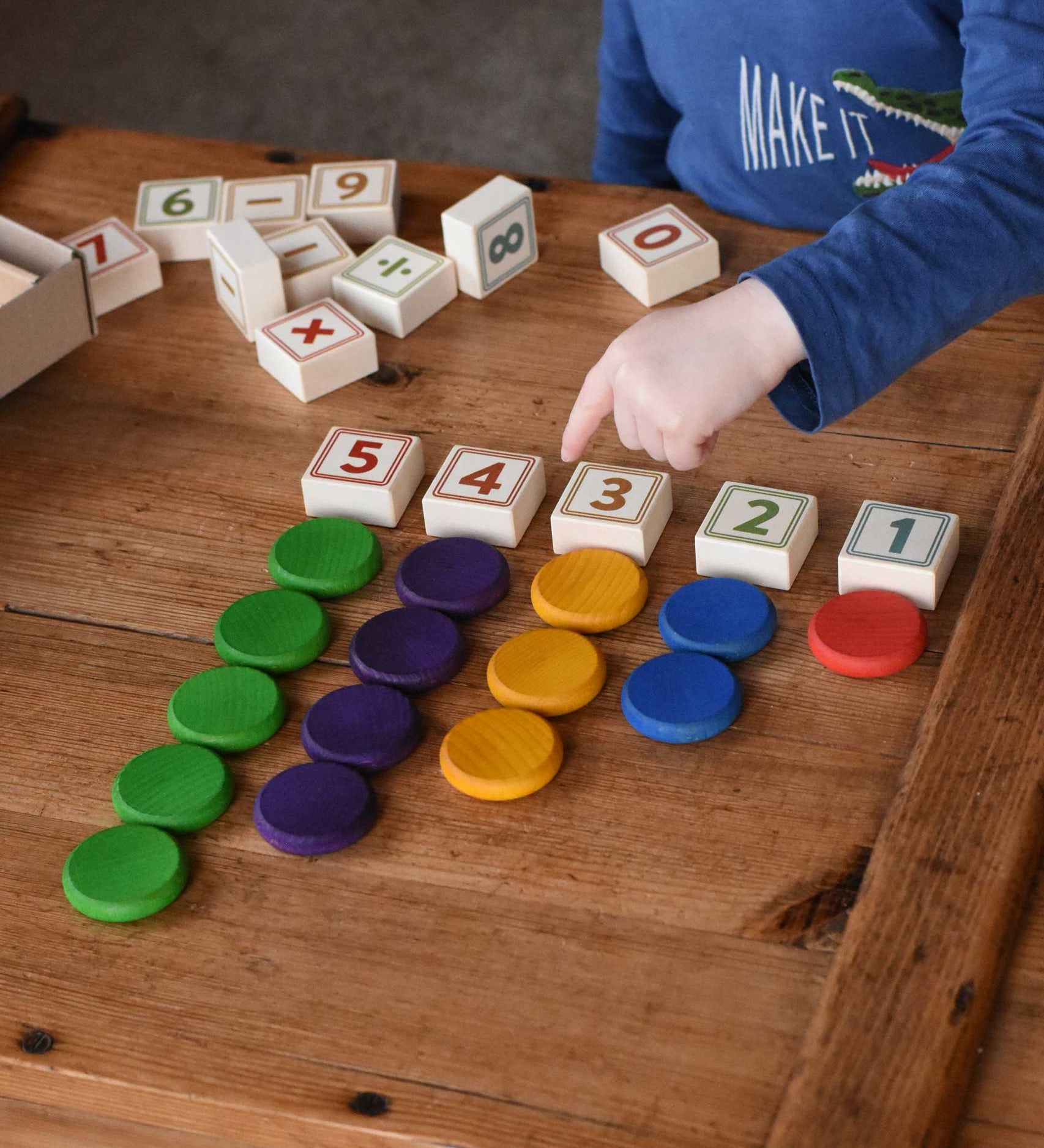 A child playing the blocks from the Bajo 40 Numbers & Symbols Blocks set. The 1 to 5 number blocks have been lined up with the corresponding number of Grapat wooden coins placed next to them. These heirloom quality toys by Bajo are part of a wide range of educational wooden toys available here at Babipur. 
