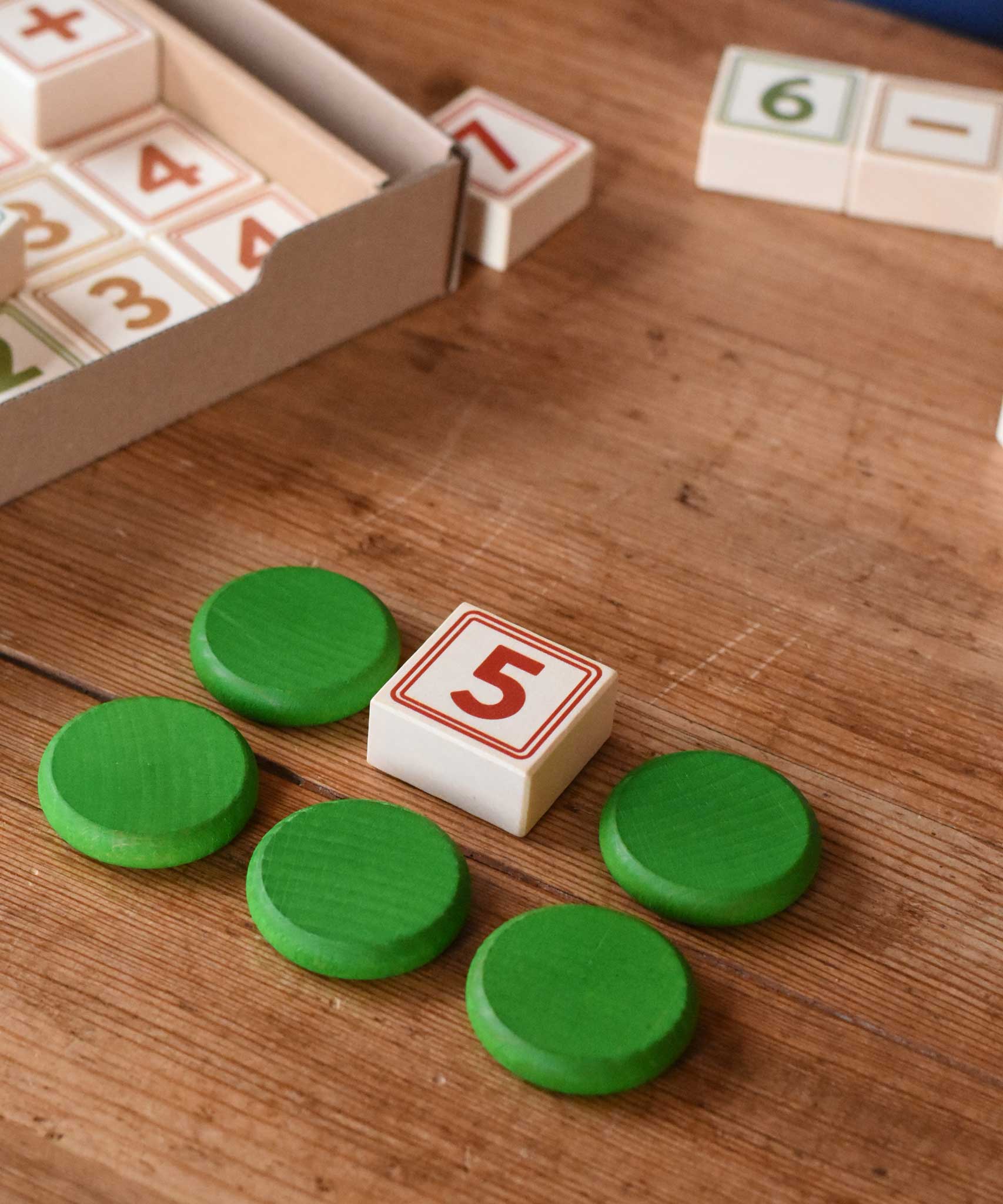 A number  block from the box of Bajo 40 Numbers & Symbols Blocks set placed on a wooden tabel next to 5 Grapat wooden coins. These heirloom quality toys by Bajo are part of a wide range of wooden toys available here at Babipur. 