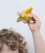 A child holding up a yellow coloured Bajo toy plane with pilot above their head. This plane is part of a wide range of Bajo wooden toy vehicles available at Babipur.