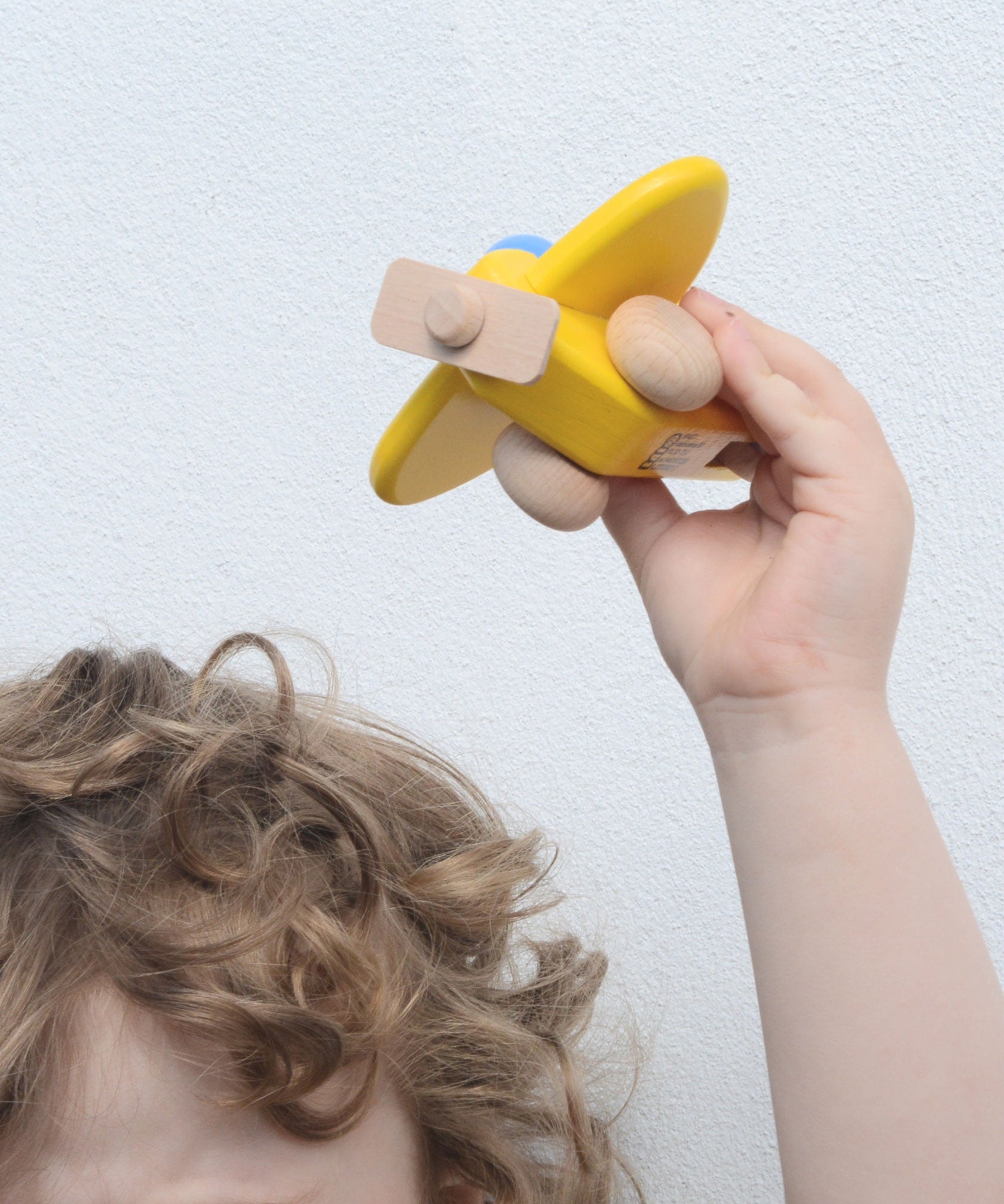 A child holding up a yellow coloured Bajo toy plane with pilot above their head. This plane is part of a wide range of Bajo wooden toy vehicles available at Babipur.