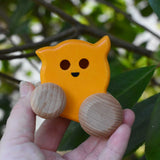 A Bajo push along eye ghost wooden toy shown in an adult's hand. The toy has a yellow painted body and natural wooden wheels. These heirloom quality toys by Bajo are part of a wide range of wooden toys available here at Babipur. 