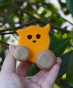 A Bajo push along eye ghost wooden toy shown in an adult's hand. The toy has a yellow painted body and natural wooden wheels. These heirloom quality toys by Bajo are part of a wide range of wooden toys available here at Babipur. 