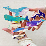 The wooden whale figures shown in an adult's hands to show scale. These heirloom quality toys by Bajo are part of a wide range of wooden toys available here at Babipur. 