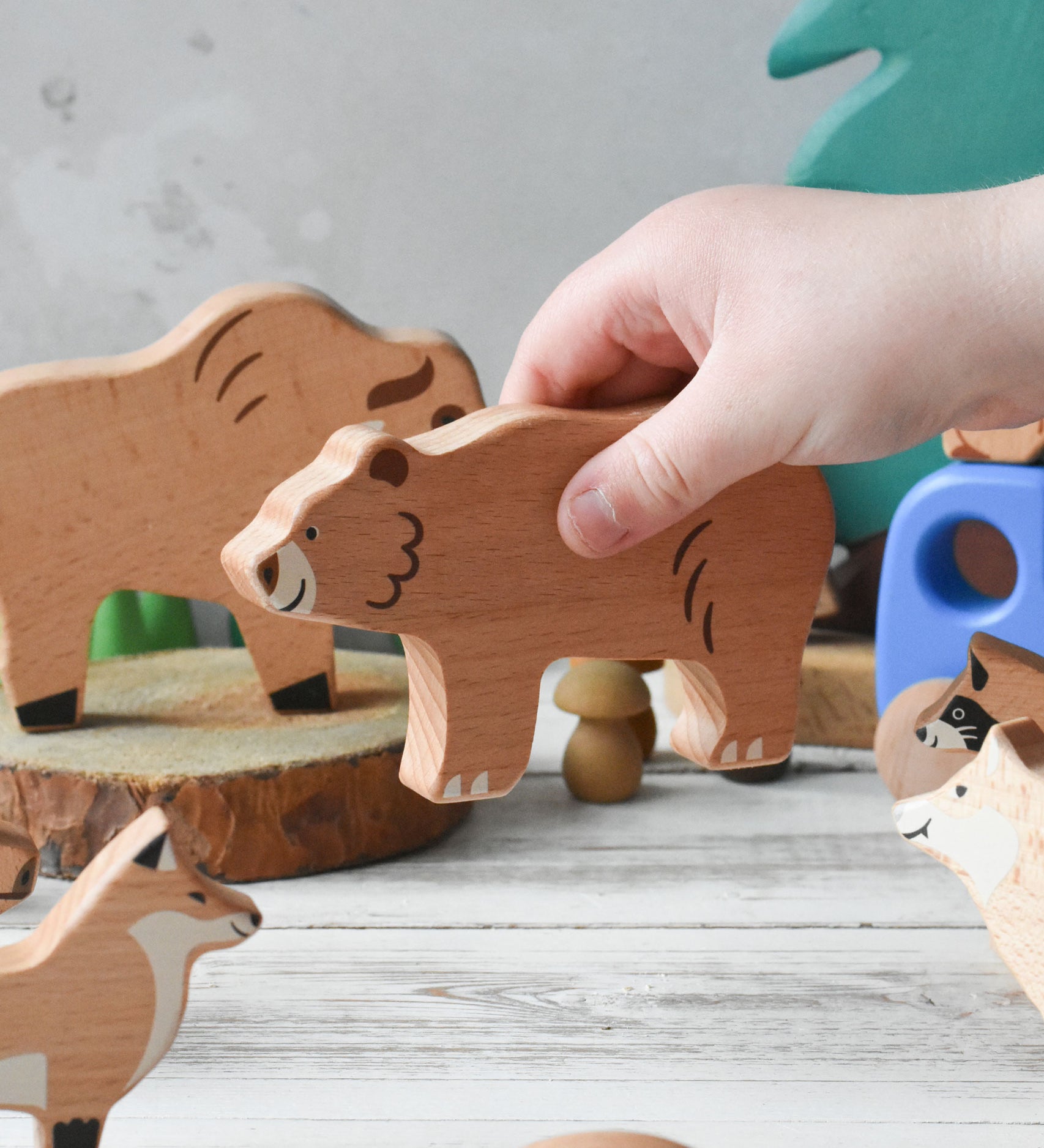 A close up of a child's hand placing the wooden bear figure from the  Bajo Wild Animals Forest Set in a small world play scene. A set of 10 wooden forest animal figures that include: Fox, Wolf, Bison, Squirrel, Hedgehog Deer, Hog, Owl, Raccoon and Bear. These heirloom quality toys by Bajo are part of a wide range of wooden toys available here at Babipur.