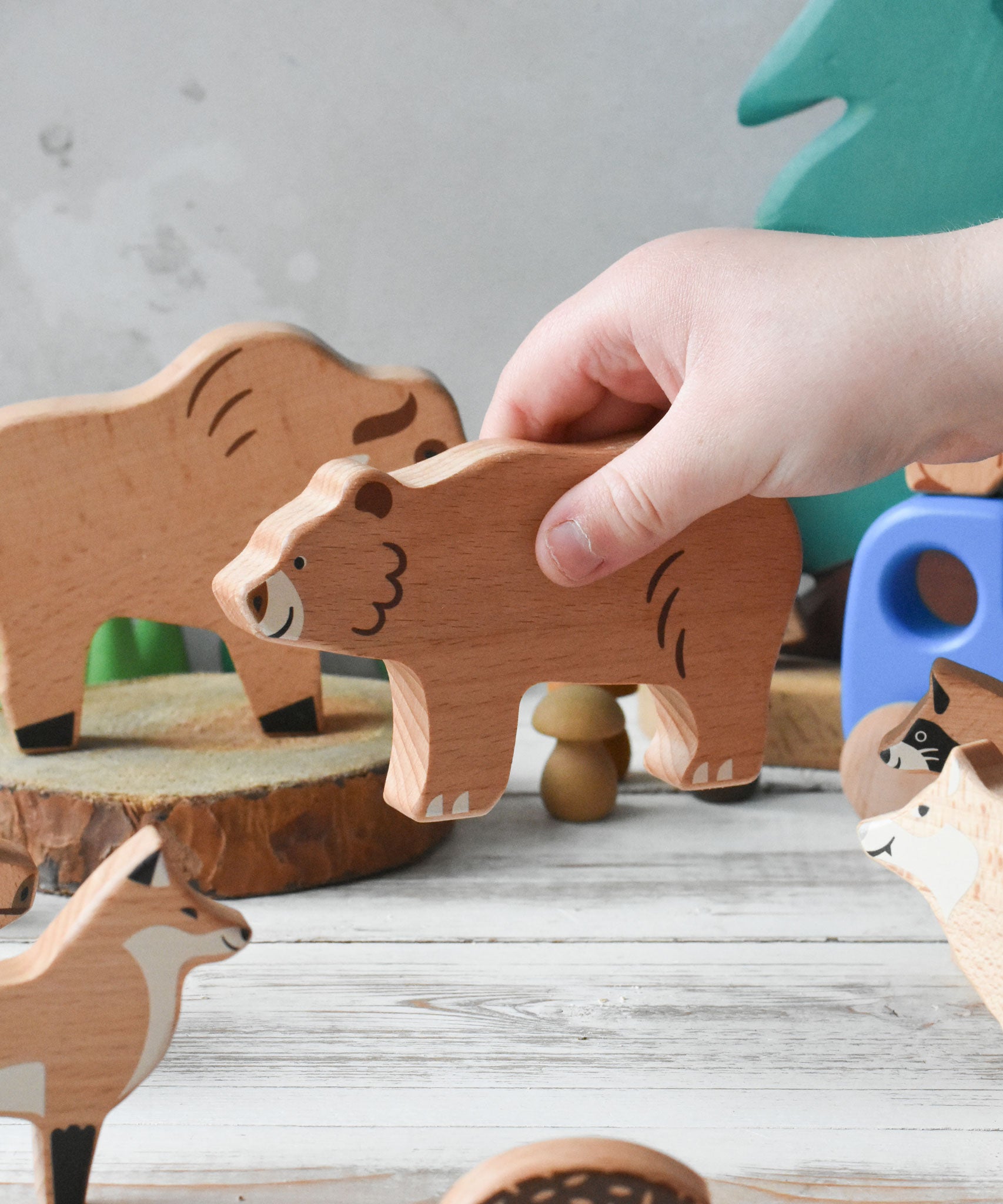 A close up of a child's hand placing the wooden bear figure from the  Bajo Wild Animals Forest Set in a small world play scene. A set of 10 wooden forest animal figures that include: Fox, Wolf, Bison, Squirrel, Hedgehog Deer, Hog, Owl, Raccoon and Bear. These heirloom quality toys by Bajo are part of a wide range of wooden toys available here at Babipur.