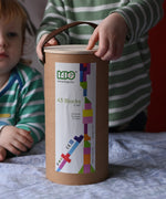 Bajo 43 wooden building blocks. 43 blocks, in 16 different shapes and 10 colours. Child lifting the lid off the sturdy cylinder shaped storage box.