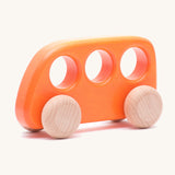 The Bajo push-along bus, a thin wooden bus-shaped toy with three 'window' holes for easy grasping. The main body is painted with non-toxic paints in orange, and the four wheels are left natural to show off the grain.