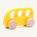 The Bajo push-along bus, a thin wooden bus-shaped toy with three 'window' holes for easy grasping. The main body is painted with non-toxic paints in yellow, and the four wheels are left natural to show off the grain.