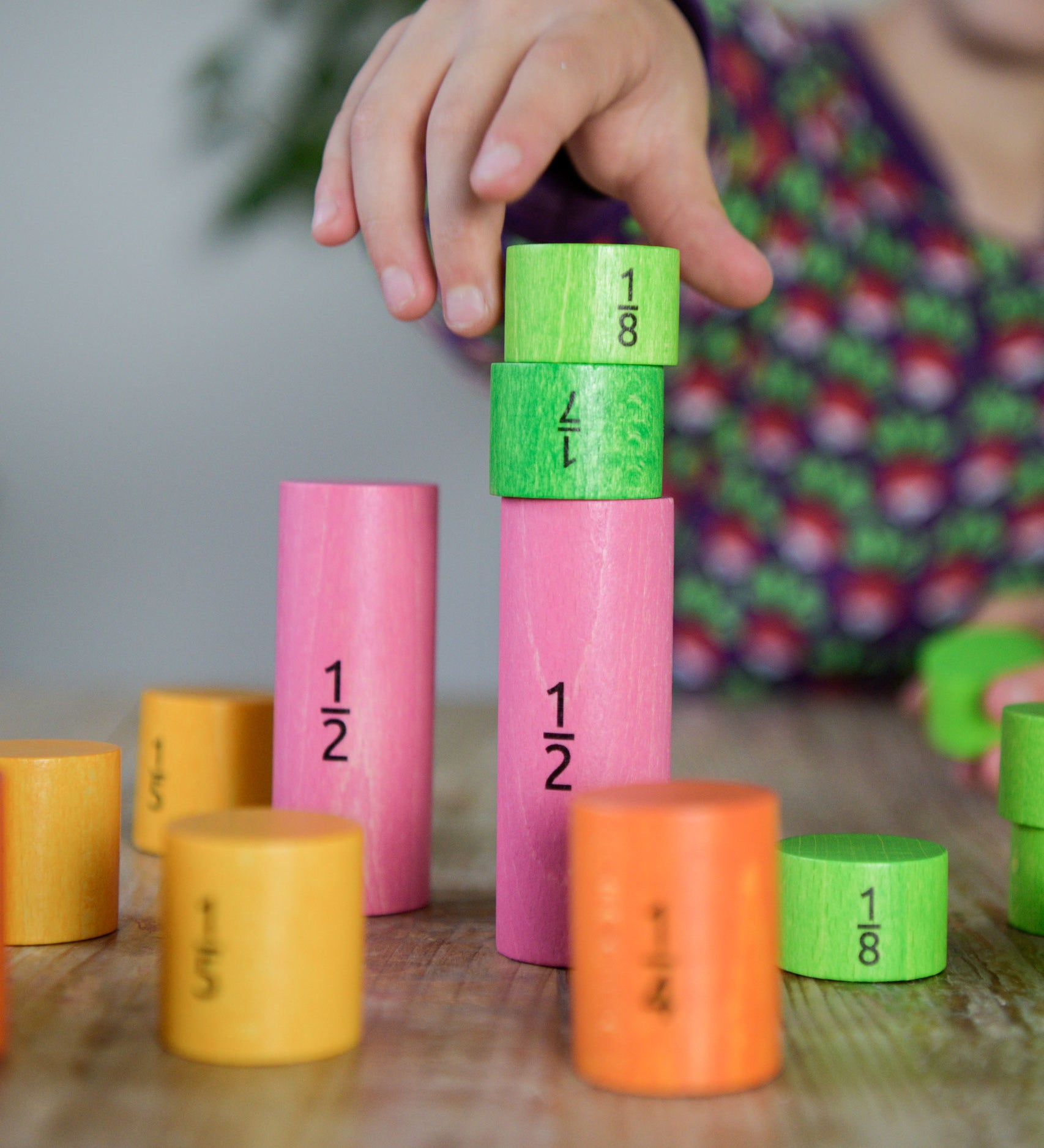 A close up of a child's hand playing with the Bajo Fractions Set. A set of coloured wooden blocks marked with fractions on each block. These heirloom quality toys by Bajo are part of a wide range of wooden toys available here at Babipur.