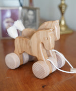 Bajo hand made wooden natural jumping horse available at Babipur. Features unpainted wheels and a toggle to pull along. Horses are standing on a wooden surface 