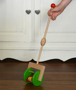 Bajo push along wooden toy rattle in a child's hand. Green rollers with coloured pieces in the middle attacked to a stick.