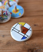 Bajo Mondrian Spinning Top placed on a wooden surface with other wooden spinning tops in the background.