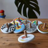 Bajo Mondrian Spinning Top spinning and blurred in motion a wooden surface with lots of other Bajo spinning tops placed in the background.