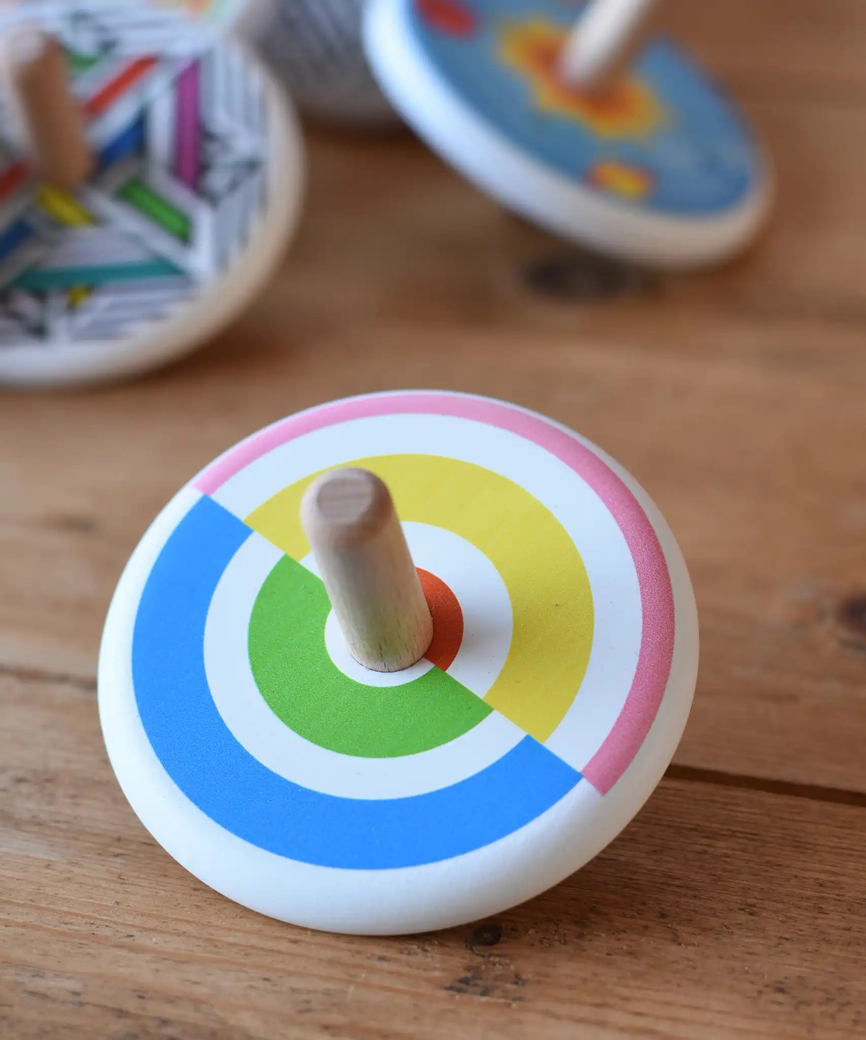 Bajo Colourful Semi Circles Spinning Top placed on a wooden surface with other wooden spinning top toys in the background.