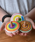 Three Bajo spinning tops in a child's hands each with a diffrent design, colourful semi circles, circles and rainbow. 