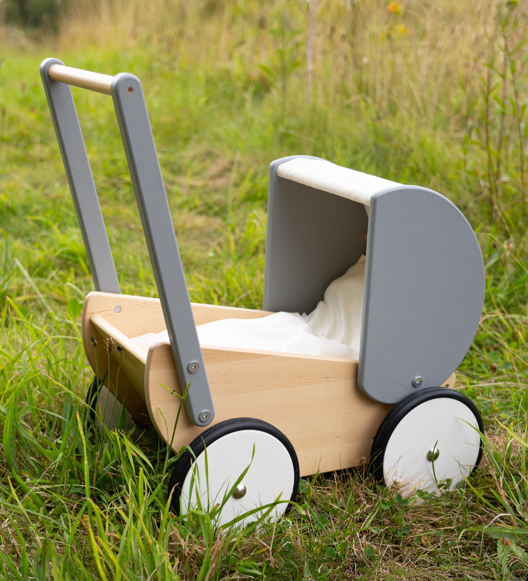 A Children's Bajo Monochrome wooden Doll's Pram placed on grass in the Babipur garden. A natural wooden and grey painted toy pram with large wheels.