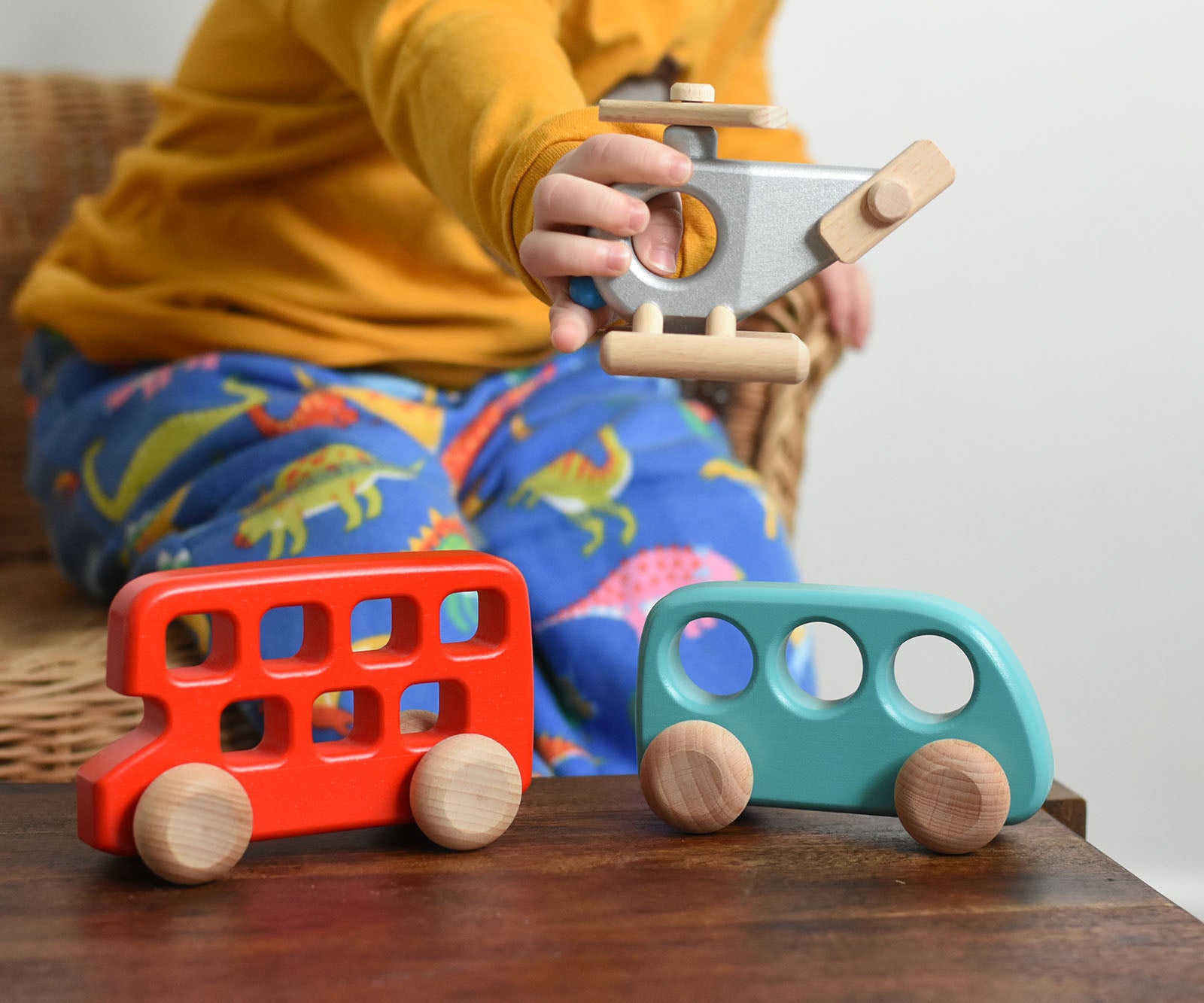 A child playing with Bajo wooden toys. The child has a wooden police helicopter in their hand with a red double decker bus and a blue bus placed on a small wooden table in front of them. These heirloom quality toys by Bajo are part of a wide range of wooden toys available here at Babipur.