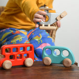 A child playing with Bajo wooden toys. The child has a wooden police helicopter in their hand with a red double decker bus and a blue bus placed on a small wooden table in front of them. These heirloom quality toys by Bajo are part of a wide range of wooden toys available here at Babipur.