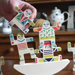 An adult's hand can be seen holding a piece from the Bajo world balance game. Three figures have been balanced on the base piece behind.