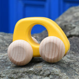 Bajo hand made wooden yellow mini style car available at Babipur. Features unpainted wheels and a cut out hole for easy grabbing. Car is sitting on a stone wall with a blue door in the background.