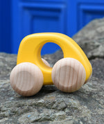 Bajo hand made wooden yellow mini style car available at Babipur. Features unpainted wheels and a cut out hole for easy grabbing. Car is sitting on a stone wall with a blue door in the background.