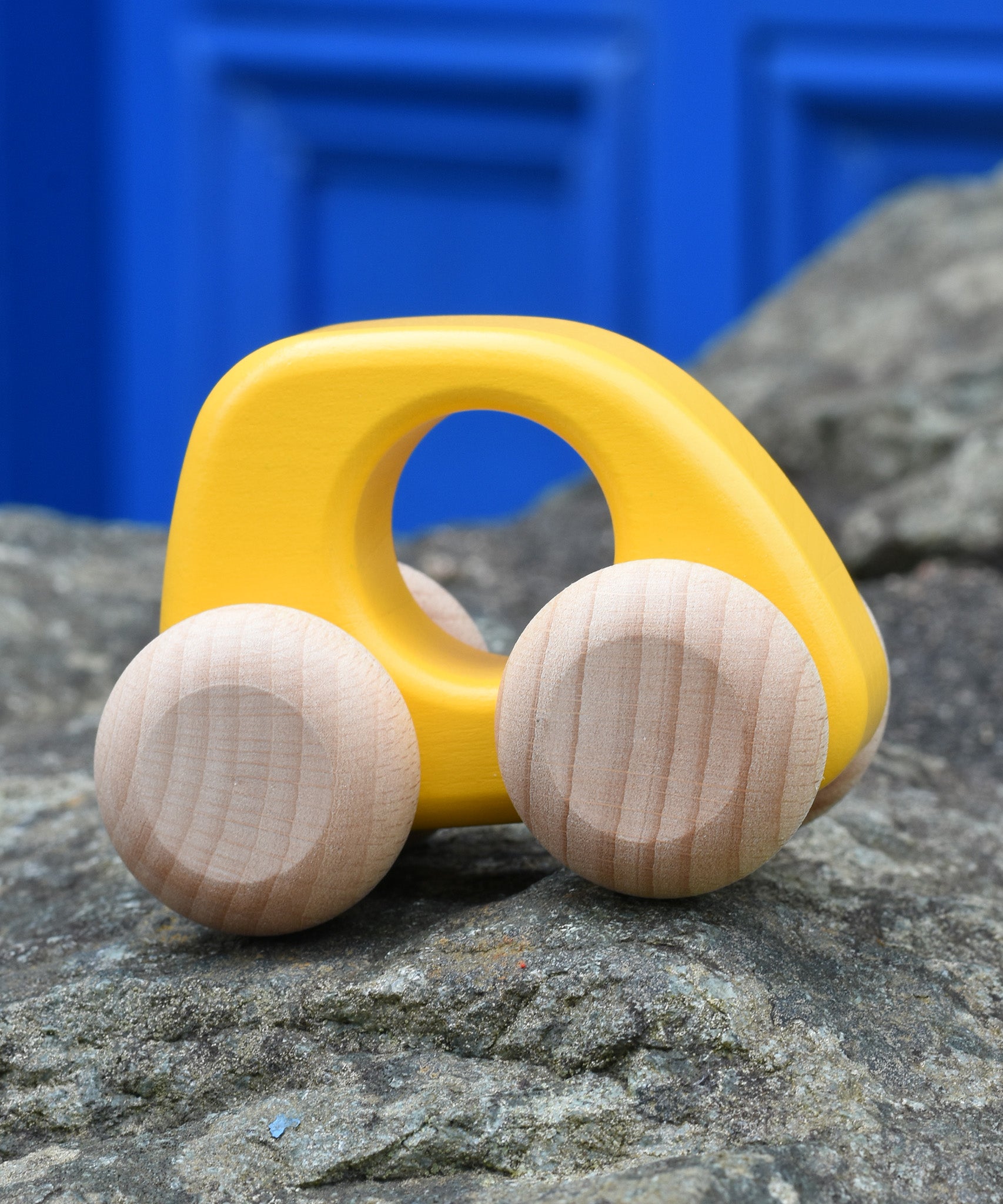 Bajo hand made wooden yellow mini style car available at Babipur. Features unpainted wheels and a cut out hole for easy grabbing. Car is sitting on a stone wall with a blue door in the background.