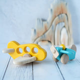 A yellow Bajo airplane with windows placed on a white wooden surface next to a blue plane with pilot. These wooden planes are part of a wide range of expertly hand crafted wooden toys here at Babipur. 