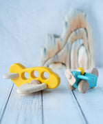 A yellow Bajo airplane with windows placed on a white wooden surface next to a blue plane with pilot. These wooden planes are part of a wide range of expertly hand crafted wooden toys here at Babipur. 