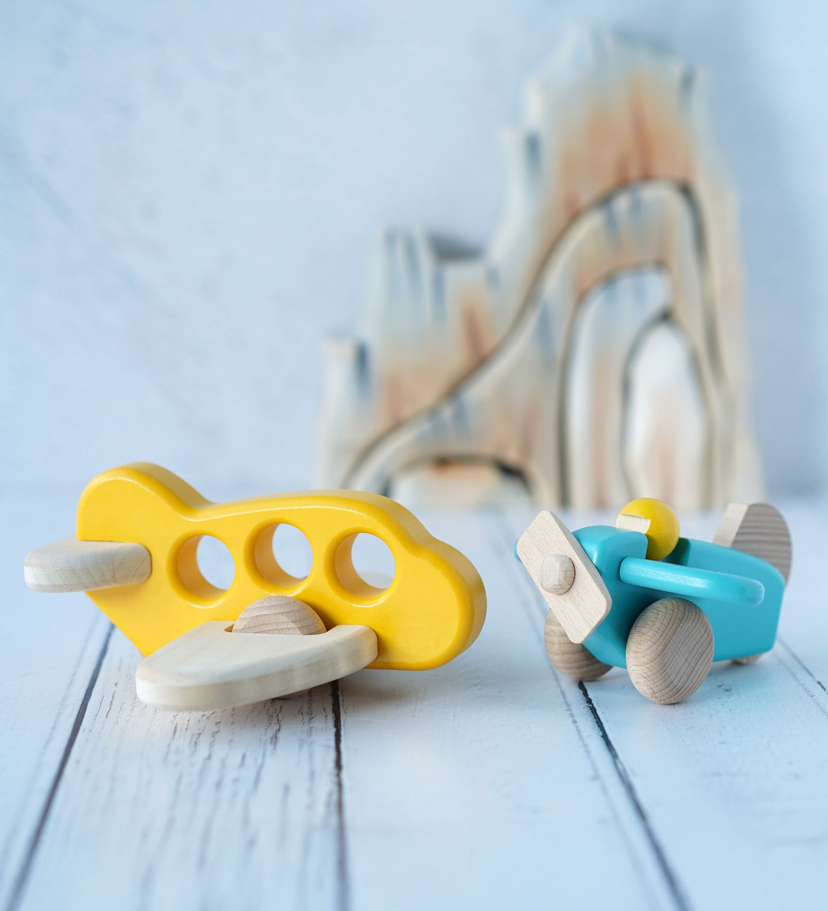 A yellow Bajo airplane with windows placed on a white wooden surface next to a blue plane with pilot. These wooden planes are part of a wide range of expertly hand crafted wooden toys here at Babipur. 