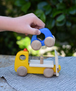 Bajo yellow breakdown lorry wooden toy with a child's hand lifting the blue car off the trailer.