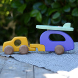 Bajo yellow car and purple camper wooden toy vehicle with green surfboard placed on a wooden surface outdoors.