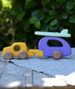 Bajo yellow car and purple camper wooden toy vehicle with green surfboard placed on a wooden surface outdoors.