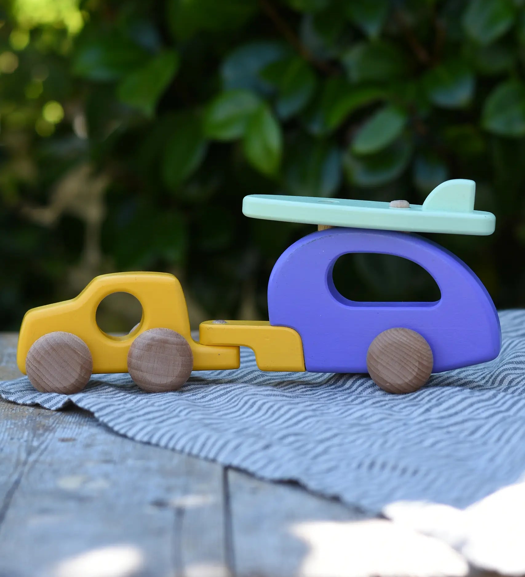 Bajo yellow car and purple camper wooden toy vehicle with green surfboard placed on a wooden surface outdoors.